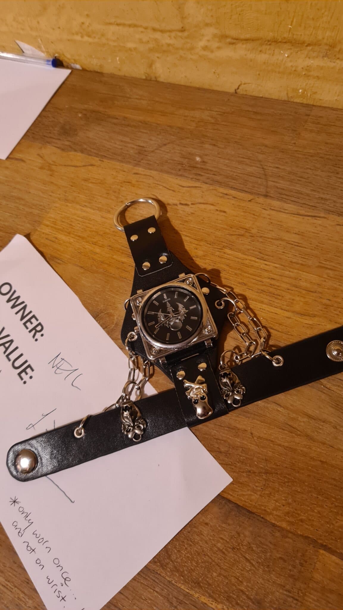 A strange gothic inspired watch on perpendicular leathers straps with added chains with asquare watch case and black dial and bird motif on top fo the dial also, ying on a piece of paper on a brown table