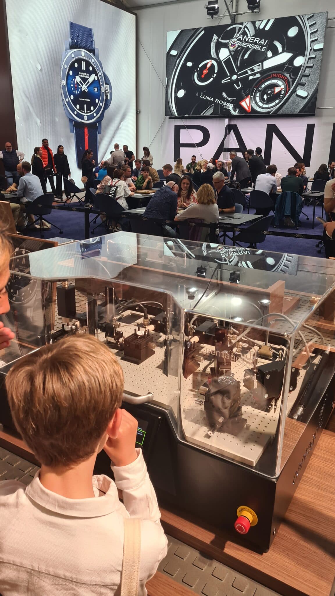 a small child with ligth brown hair and white shirt on looking at a large watch testing machine at the panerai stand, with tables and gorups ofpeople looking at watches in the background and large watch pictures visible on the walls behind them