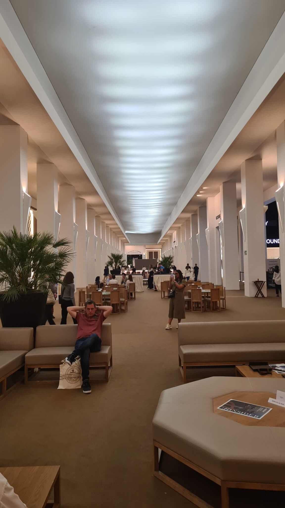 a large hallway filled with seats and plants with white columns either side and abeie roof, with people sitting and standing around