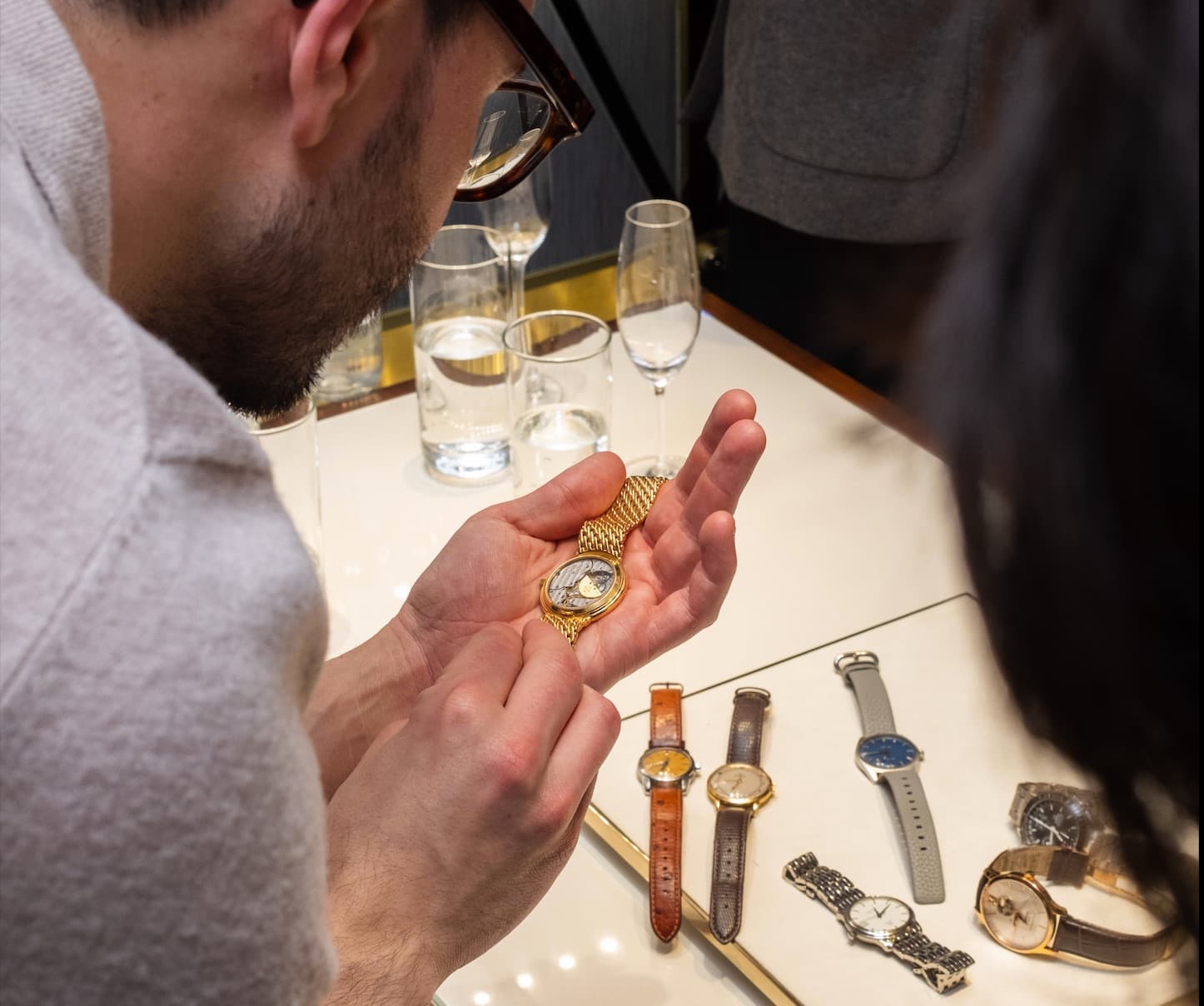 a man in a grey t-shirt with glasses looking closely at a watch in his hands with more watches on the table in front of him