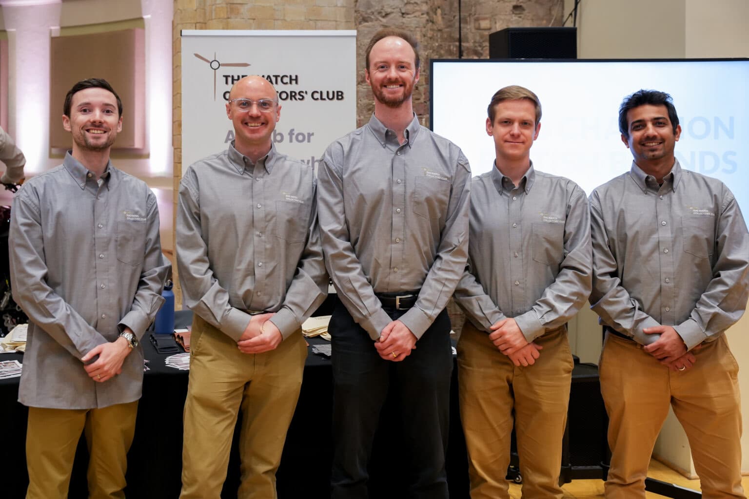 the volunteer team for The Manchester Watch Show from The Watch Collectors Club, wearing grey club shirts standing in a line with hands clasped in front, five men