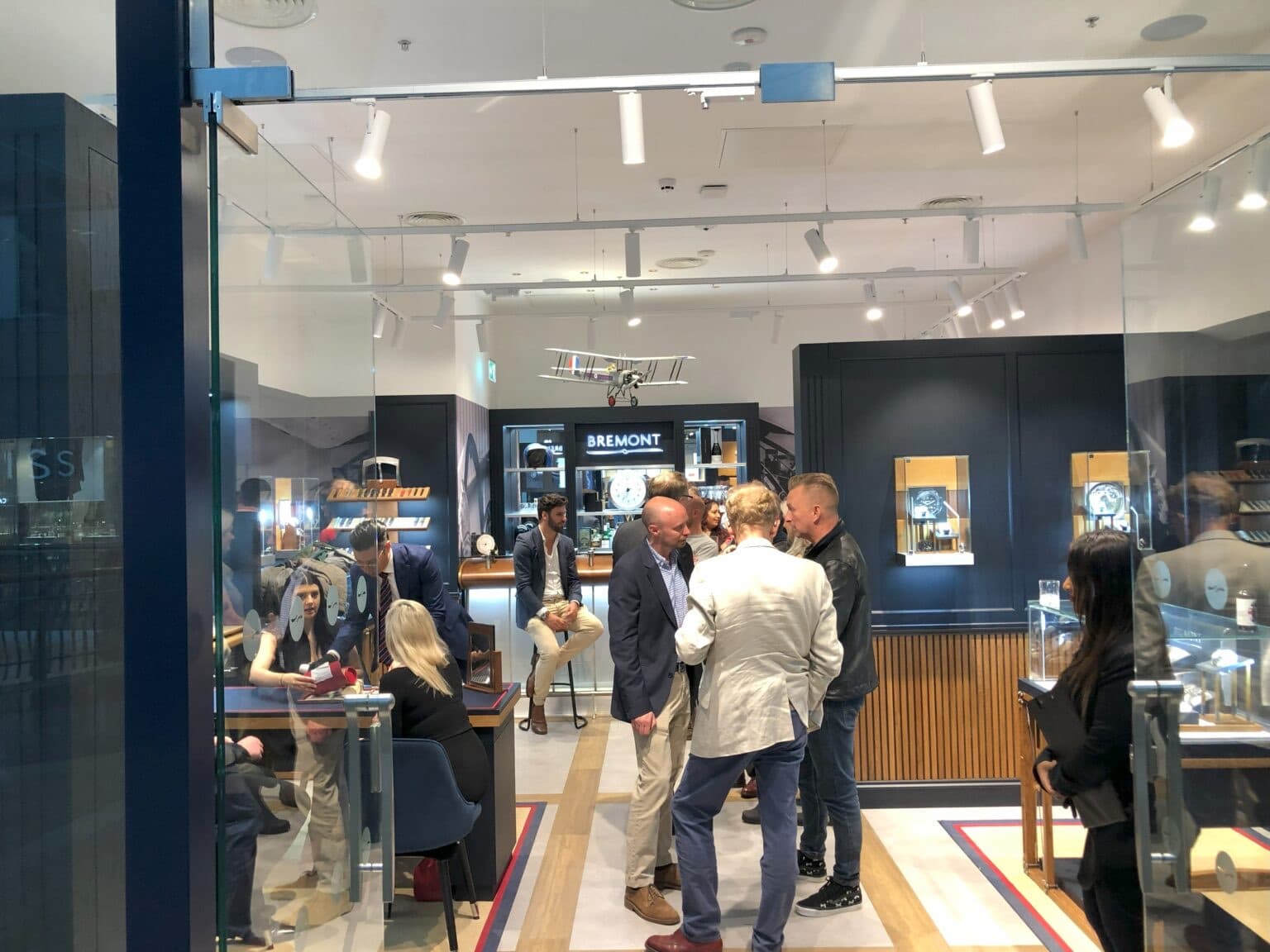 picture looking into a Bremont watch boutique with people talking in the middle of the store, people sitting at a desk on the left chatting about watches, and watch memorabilia surrounding them on the walls and watches in cabinet, blue overall theme and glass shopfront and doors