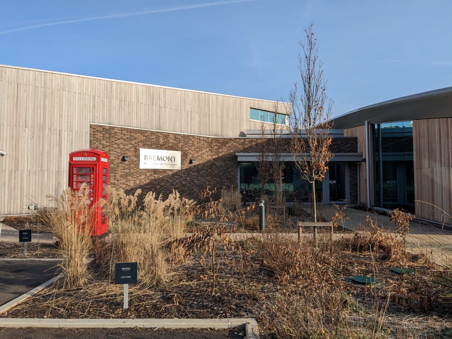 enrance to bremont factory with sign visibile and bushes and plants in the foreground, blue sky above, red phone box off to the right