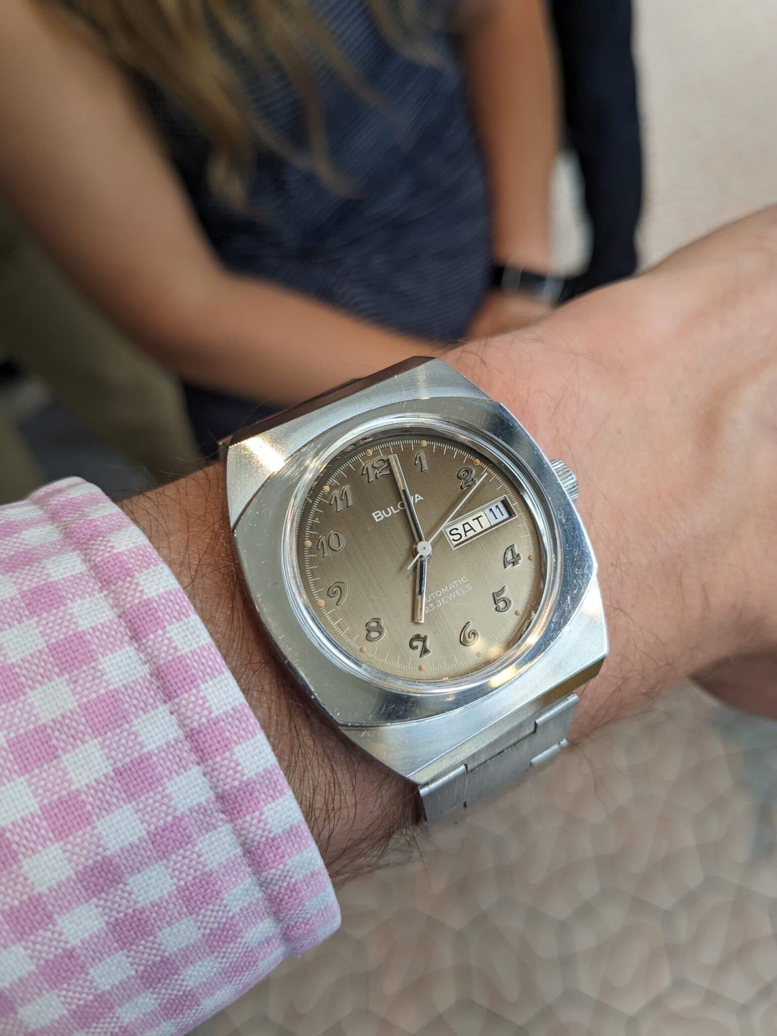 An unusual square case Bulova watch with rounded corners and coffee coloured dial which fades to lighter at the bottom on a wrist next to a pink shirt