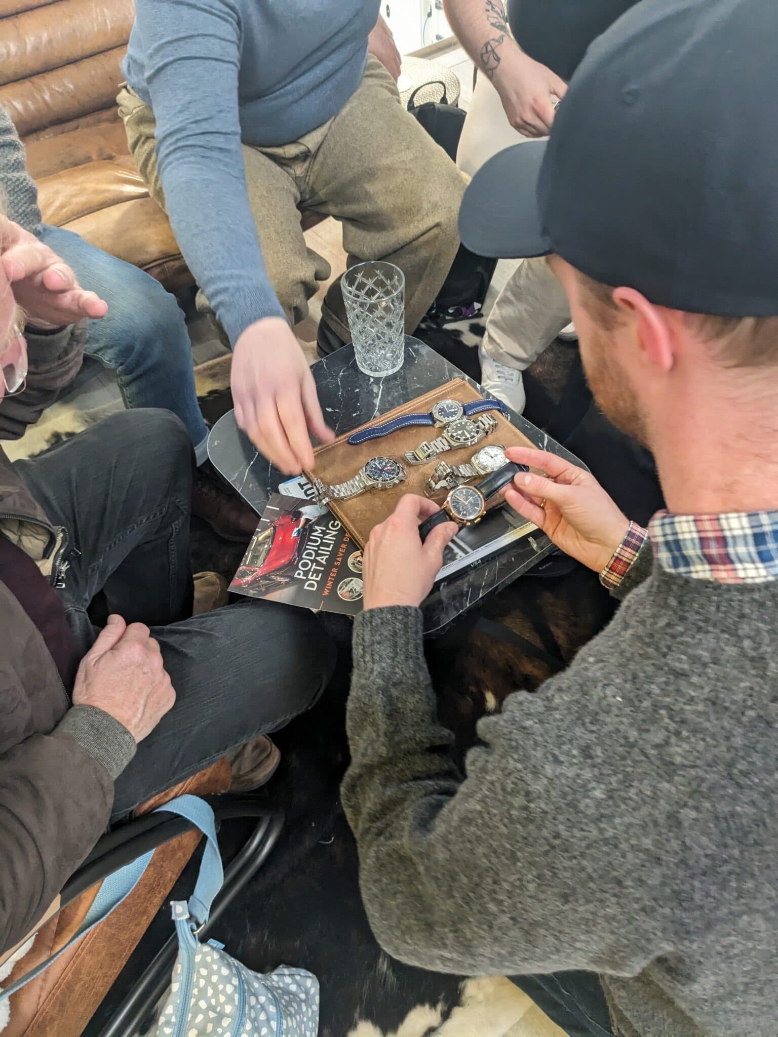 a group of people round a low table covered in watches with some people reaching out to pick up the watches, all wearing a variety of jumpers and hats