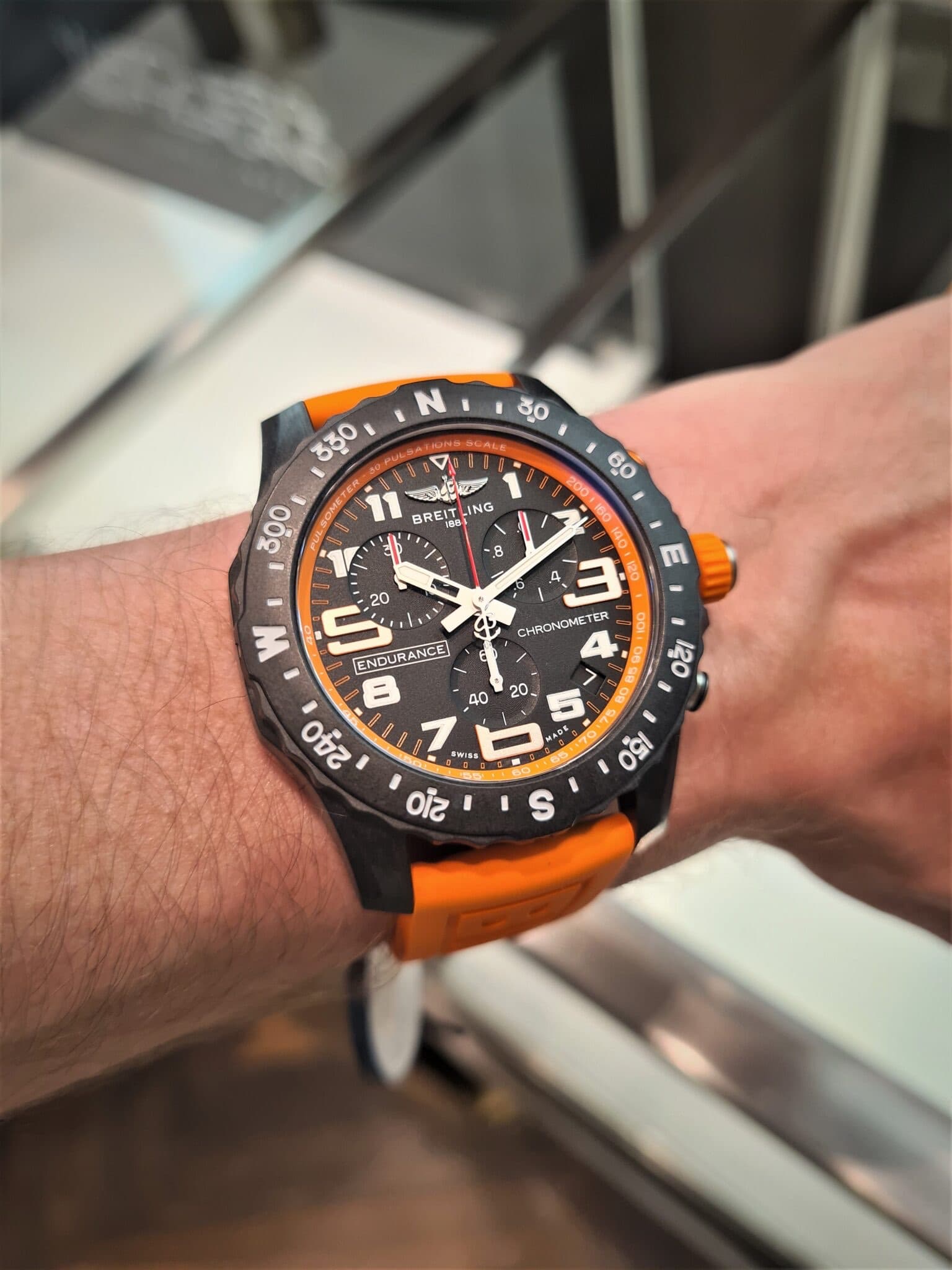 a black grey and organe sports watch from Breitling with white numbers and hands and an orange strap