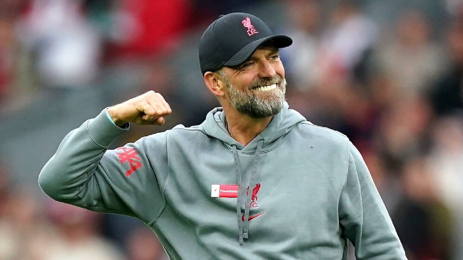 Man in grey tracksuit top with black hat holding fist clenched above his shoulder and smiling in celebration jurgen klopp with beard and bright teeth