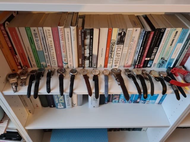 A group of watches arranges on a book shwlf with their straps hanging over the edge, with book spines facing outwards behind them