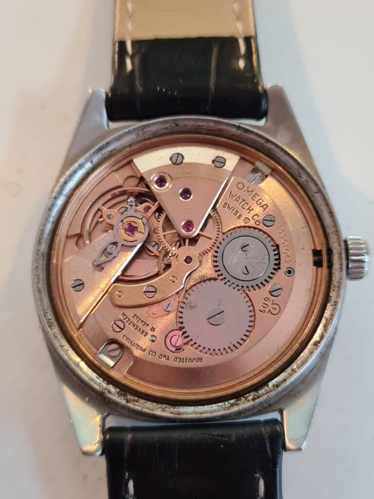 A polished brass Omega movement with steel wheels and brass bridges, all a rose gold colour in a steel case.