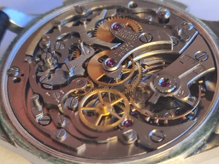 a round open watch case showing the mechanism or movement of this chronograpg, with steel and brass wheels and bridges, screws, levers and more. the escapement is visible and the chronograph mechanism wheels also