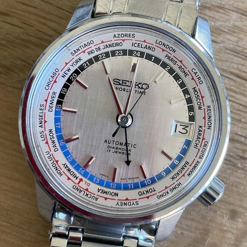 seiko worldtimer for the 1964 olympic games with silver dial with blue and blakc edge, red highlights and black cities around the edge of the dial in a steel case and bracelet with a 5 o'clock crown