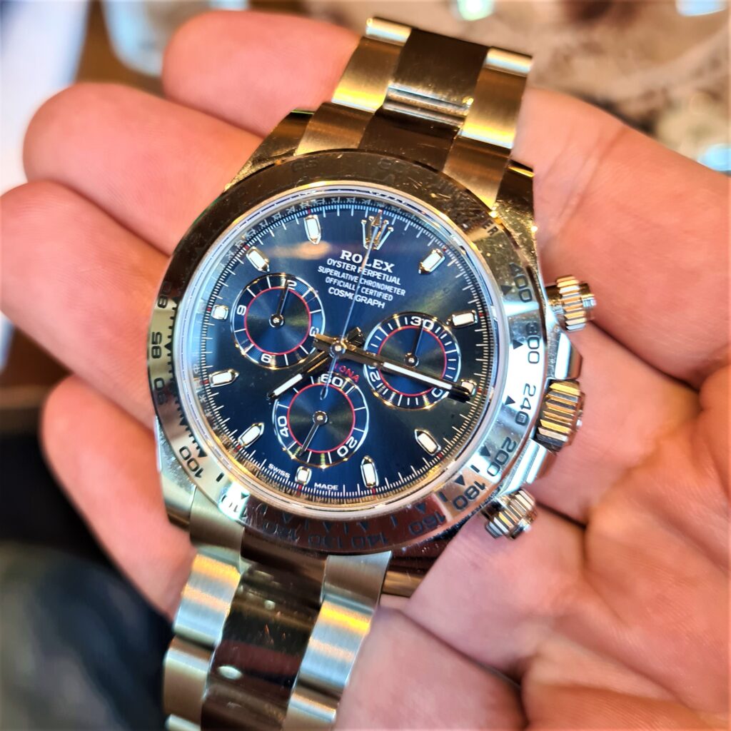 Shiny white gold Rolex Daytona watch with dark blue dial held in someone's hand with red accents