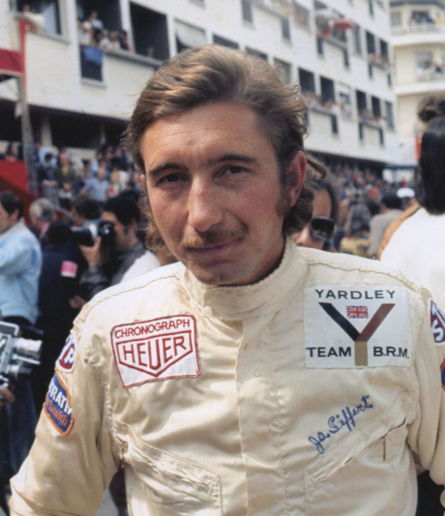 a picture of jo siffert, a white man in his late 20s win a white racing overals with sponsor logos clearly visible, in pit lane with people behind