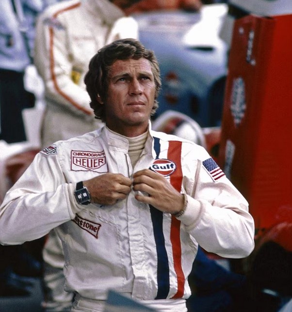steve mcqueen in his chacter outfit for le mans, a white racing overalls with blue and red gulf stripe and sponsors patches plus Moncao wristwatch on his right wrist