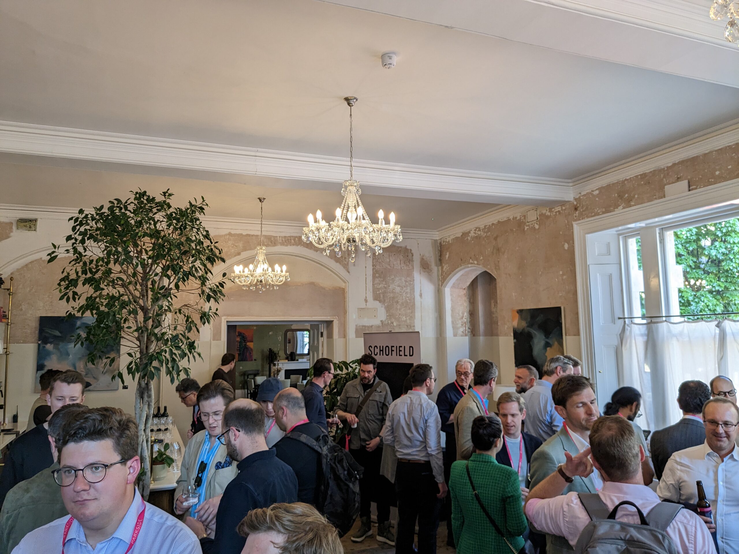 a room full of people talking with large plants, overhead chanedliers, large brigh windows, and bar on the left hand side