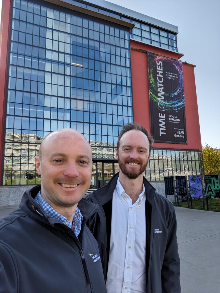 two men in grey jackets smiling in front of a large conference centre with glas facing wall and large show poster on the right saying time to watches