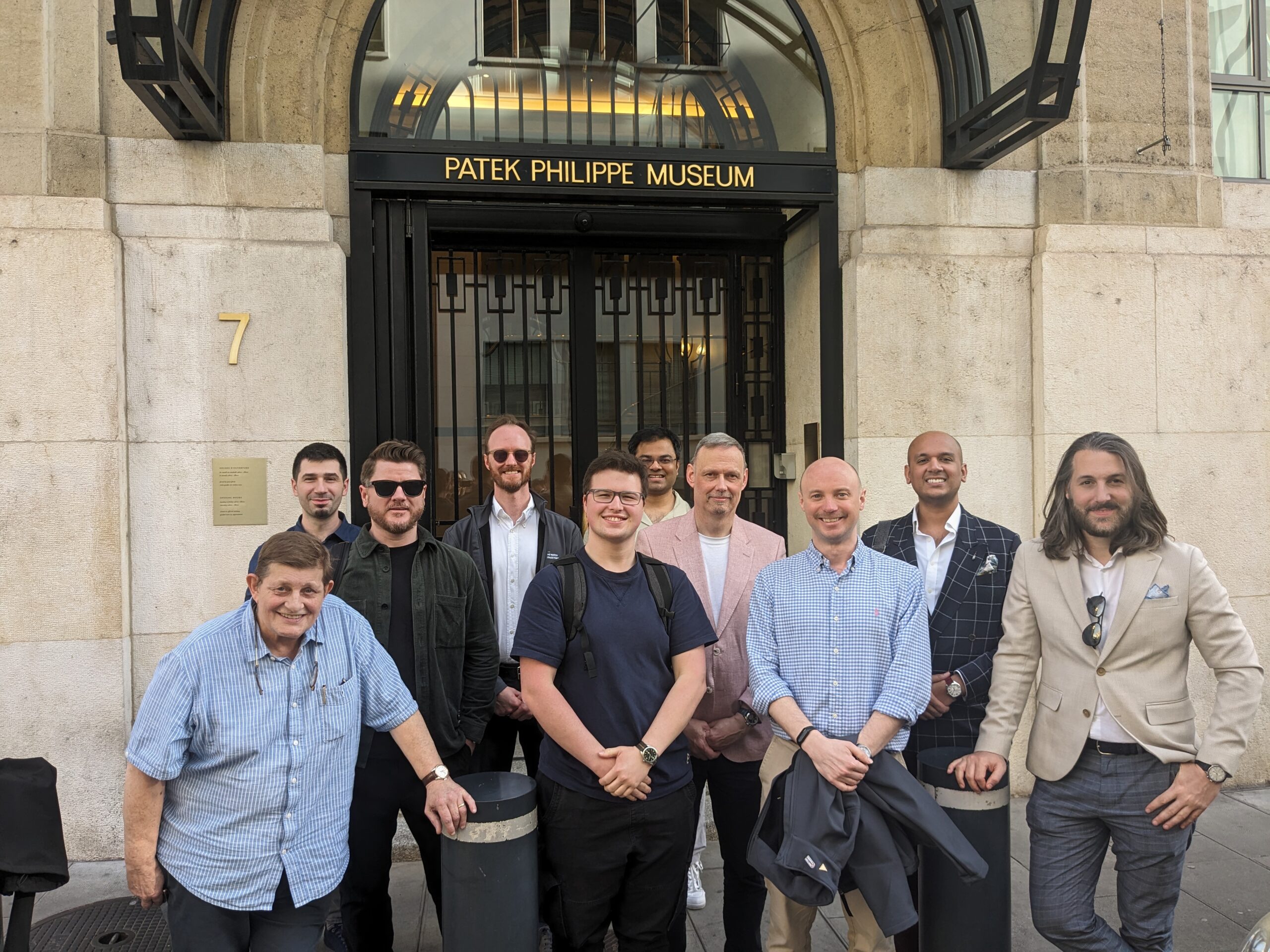 group of members of The Watch Collectors' Club in a range of outfits standing on teh steps outside the Patek PHilippe museum with beige stone walls and black iron gates and a gold sign with the name above the door