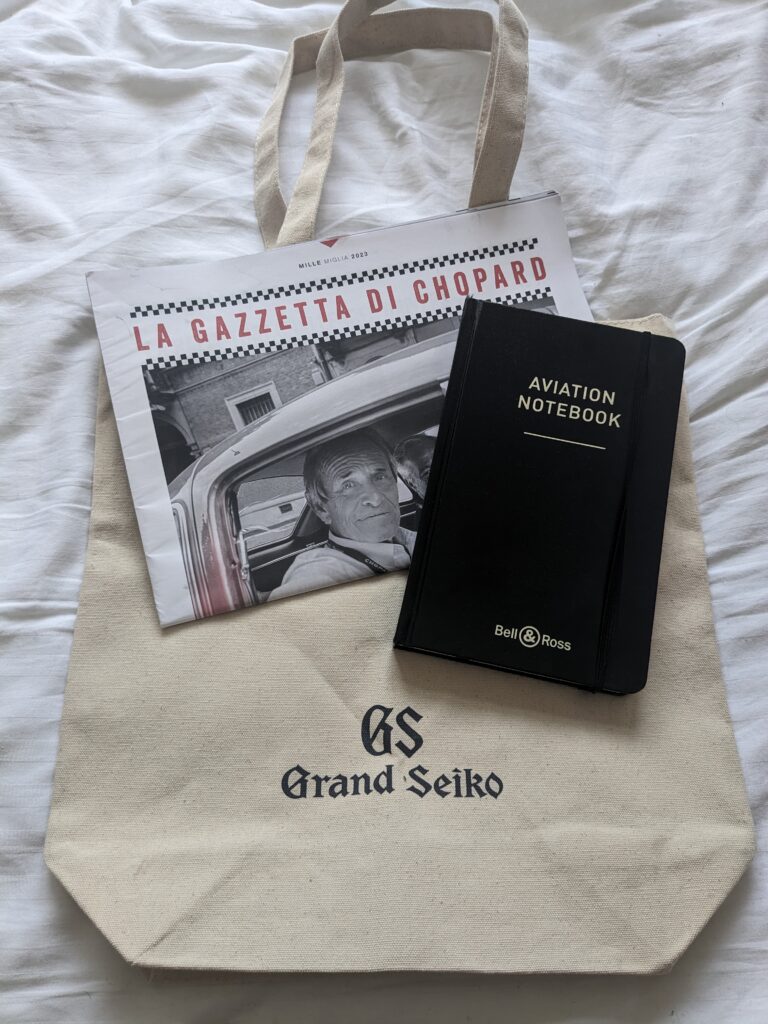 a tote beige Grand Seiko Tote bag, a black bell and ross labelled notebook and a newspaper called the cazette du chopard all lying on a white bedsheet