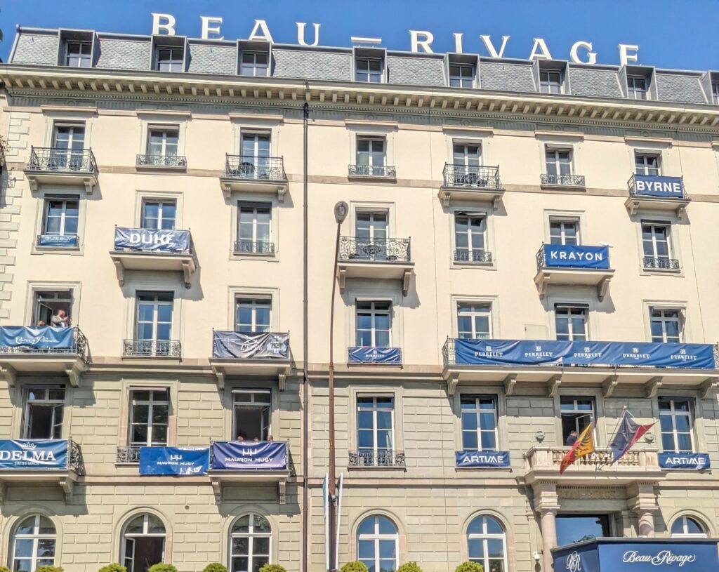 large hotel building with beige stone and balconies with blue banners flying showing watch brand names and the name of the hotel in a large sign on the roof saying beau rivage