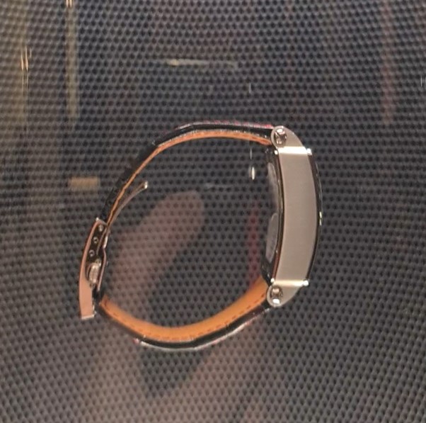 Side view of a watch with a thick rectangular case in steel with an orange and black leather strap against a grey background