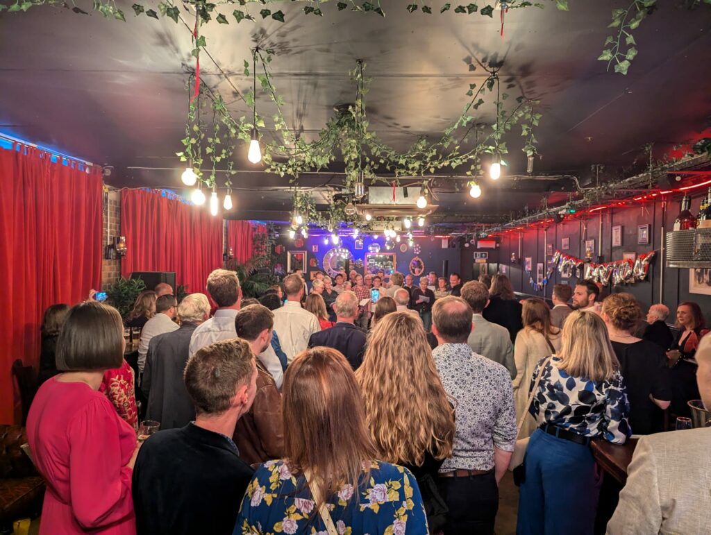 large group of people seen from behind watching a small choir in a large nightclub type room with dark ceiling