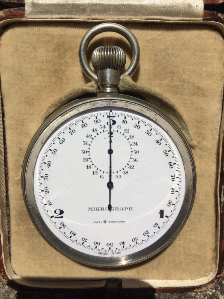 a large 1930s mikrograph pocket watch style chronograph with bright white enamel dial and small nuimbers around the ourtside with think black hands with crown and pusher on top restingin beige cloth case