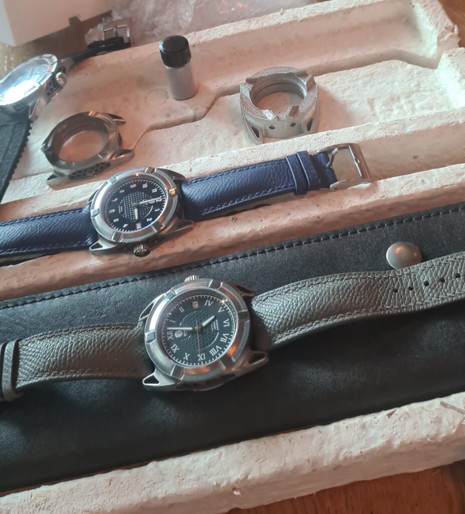 apiar invenire watches on a table on top of a black leather watch pouch and mycellium box, with case prototypes in the background, the watches have green and blue dials and leather straps